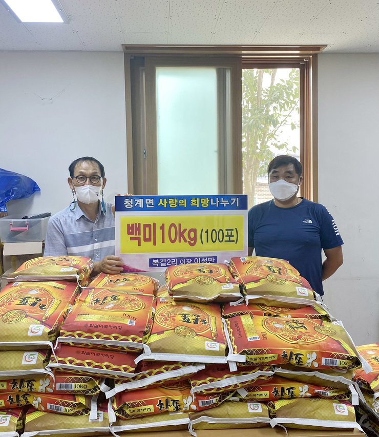 청계면 사랑의 희망나누기 백미 10kg (100포) 복길2리 이장 이성만 이라고 적힌 배너를 들고 후원품인 백미를 앞에 쌓아두고 기념촬영을 하고 있다.