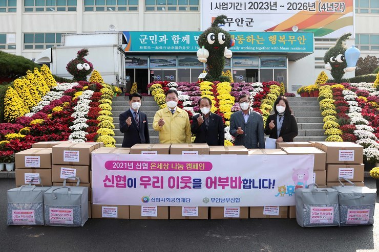 후원품을 앞에 두고 기념사진을 촬영하고 있다. 후원품 앞으로 '2021년 신협 온세상 나눔 캠페인 신협이 우리 이웃을 어부바합니다! 신협사회공헌재단 무안남부신협'이라고 적힌 현수막이 걸려있다