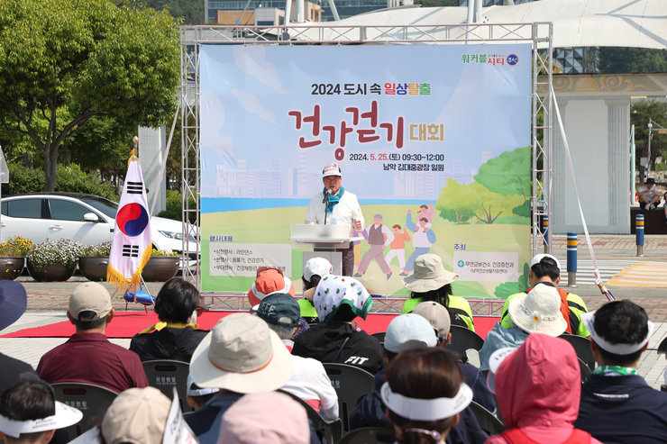 2024 도시 속 일상탈출 건강걷기 대회 2024.5.25(토) 09:30~12:00 남악 김대중광장 일원 판넬이 세워진 포토존에서 김산 시장이 앉아있는 참가 주민들을 향해 무언가를 말하고 있다.