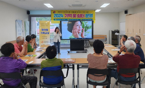 중앙에 피부를 두드리는 중년여성의 영상이 나오는 스탠드 TV를 중심으로 ㄷ자로 배치 된 테이블에 앉아 강사를 보며 따라하는 수강생들의 모습